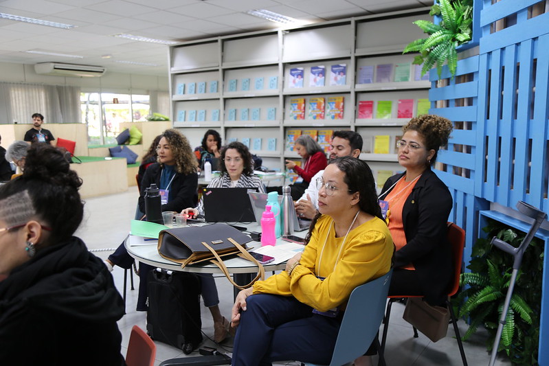 Foto com cinco pessoas sentadas ao redor de uma mesa redonda. São quatro mulheres e um homem, todos eles com olhares direcionados a um só local. 