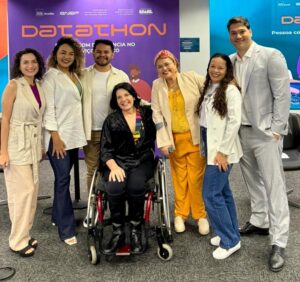 Na foto, há cinco mulheres e dois homens com roupas coloridas. Todos estão de pé e pousam para a câmera, exceto uma das mulheres, que está sentada em uma cadeira de rodas.