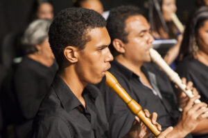 Foto com dois homens de pele negra e camisa preta tocando flauta com outras pessoas ao fundo. 