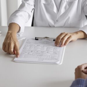 Mãos humanas sobre um papel em uma prancheta com anotações e uma mesa. A mesa e as vestimentas da mão são brancas. 